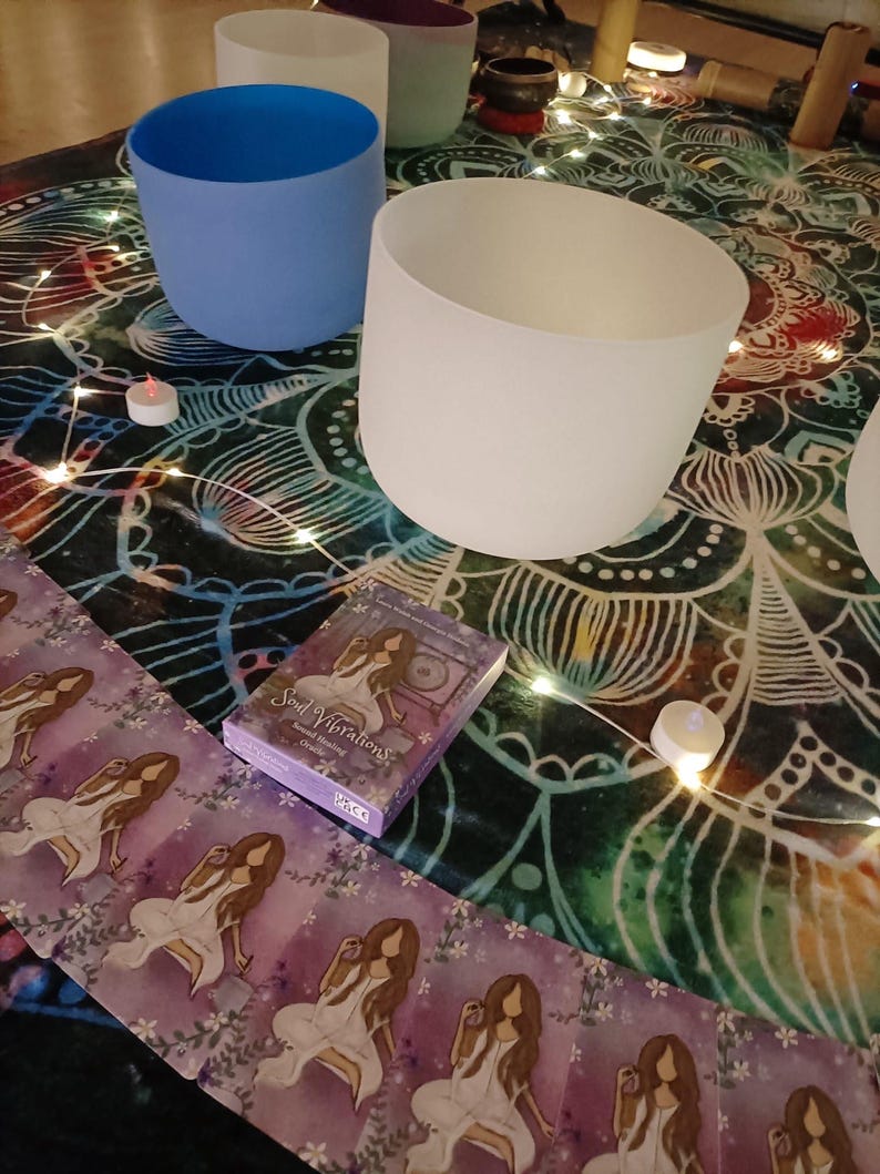 May include: A collection of frosted glass singing bowls in various colors, including white, blue, and purple, arranged on a patterned cloth. A deck of "Soul Vibrations" oracle cards and small candles are also present, suggesting a spiritual or wellness theme.