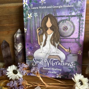 May include: A purple and white oracle card deck titled "Soul Vibrations" with the text "Sound Healing Oracle." The cover art features an illustration of a person in white clothing, a gong, and flowers. Amethyst crystals and flowers surround the deck.
