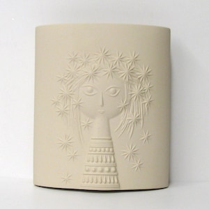May include: A beige ceramic vase with a raised relief design of a woman's face and a starburst crown. The vase is rectangular with rounded edges and stands on a white surface.