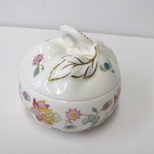 May include: A white ceramic trinket box with a lid, featuring a floral pattern in pink, yellow, and blue. The lid has a leaf-shaped handle with gold accents. The box is round and sits on a white surface.