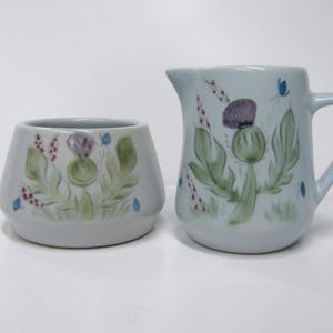 May include: A set of two ceramic creamer and sugar bowl with a thistle design. The creamer is white with a blue and green thistle design. The sugar bowl is white with a blue and green thistle design.