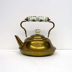 May include: A vintage brass teapot with a curved spout and a decorative handle. The handle is white with blue floral designs. The lid has a white knob. The teapot is on a white background.