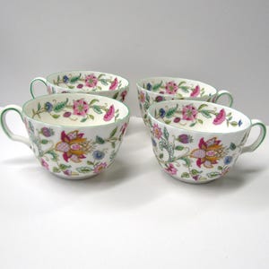 May include: Four white teacups with a floral pattern in shades of pink, yellow, green, and blue. The cups have a green rim and are decorated with a delicate floral design.