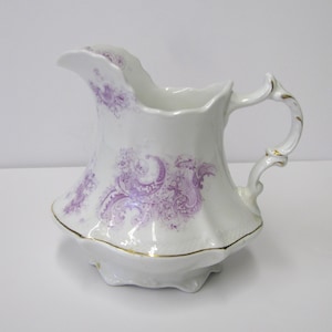 Peut inclure: Pichet en céramique blanche avec une base évasée et une anse incurvée. Le pichet présente un motif floral violet et une bordure dorée. Un objet décoratif pour servir des boissons.