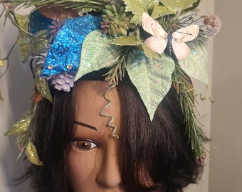 May include: A green, blue, and white glittery hat with a butterfly accent. The hat is made of various materials, including leaves, pine needles, and glitter. It is worn on a mannequin with dark hair.