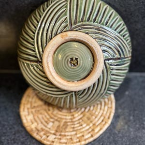 May include: A green ceramic bowl with a textured, carved design, resting on a woven coaster. The bowl has a circular base with a lighter-colored rim and a central design. The coaster is made of woven natural fibers.