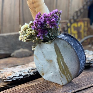 May include: A small, white ceramic vase with a black rim and a textured surface. The vase is decorated with streaks of yellow glaze and holds a bouquet of dried flowers, including purple statice and white baby's breath. A single feather is visible behind the flowers.