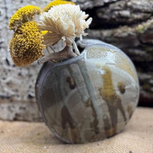Peut inclure: Un vase en céramique fait à la main, de forme ronde et avec une ouverture unique, présentant un mélange de tons terreux. Le vase contient des fleurs séchées dans des tons de jaune et de crème, créant une présentation naturelle et artistique. Le vase mesure environ 15 cm de diamètre.