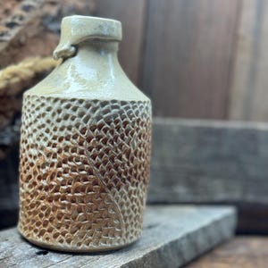 May include: A handcrafted ceramic bottle with a textured, patterned surface. The bottle has a light brown and beige color scheme, with a detailed design on the body. The bottle is displayed on a wooden surface, showcasing its rustic aesthetic.