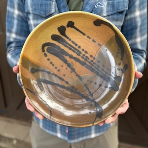 May include: A handmade ceramic serving bowl with a wide rim. The bowl features a brown and tan base with black paint drips and splatters across the surface. The bowl is round and appears to be food safe.