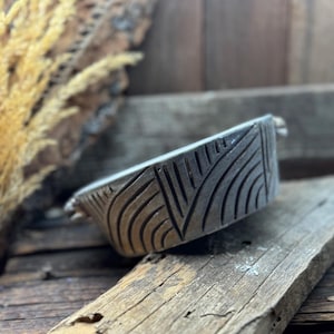 May include: A small, gray ceramic bowl with a geometric design. The bowl has a rounded shape and a slightly raised rim. The design is made up of lines and curves that create a modern and abstract look.