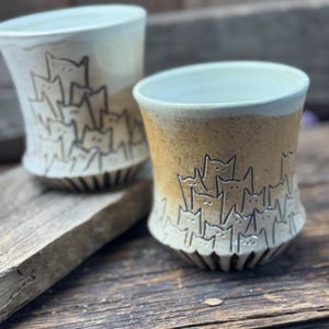 May include: Two ceramic mugs with a white glaze and a brown glaze. The mugs have a design of a stack of cats with their faces peeking out.