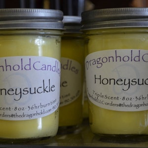 May include: Several Honeysuckle scented candles in glass jars with silver lids. Each candle has a label that reads "dragonhold Candles Honeysuckle". The candles are 8 oz and have a 36-hour burn time.