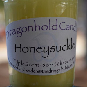 May include: A jar candle with a pale yellow wax. The label reads "dragonhold Candle Honeysuckle Triple Scent - 8 oz - 36hr burn time". The candle sits on a black lid.