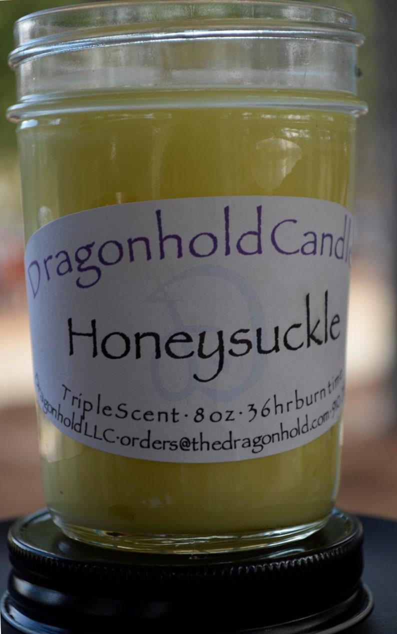 May include: A jar candle with a pale yellow wax. The label reads "dragonhold Candle Honeysuckle Triple Scent - 8 oz - 36hr burn time". The candle sits on a black lid.