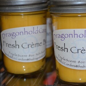 Könnte beinhalten: Nahaufnahme von mehreren Glas-Kerzen im Glas mit silbernen Deckeln und gelbem Wachs. Jedes Glas hat ein Etikett mit der Aufschrift "Dragonhold Candles Fresh Crème Brûlée". Die Kerzen sind 227g und haben eine Brenndauer von 36 Stunden.