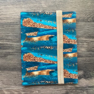 May include: A blue and gold fabric notebook cover with a gold elastic closure. The cover features a pattern of abstract brushstrokes and gold glitter.