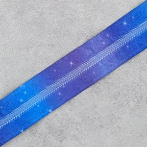 May include: A blue and purple zipper with a galaxy print and silver teeth. The zipper is lying on a gray surface.