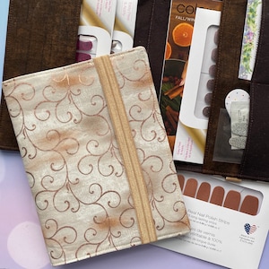 May include: A brown fabric organizer with a gold elastic band closure. The organizer has multiple pockets for storing nail polish strips. The organizer is decorated with a white and gold floral pattern. The text "Real Nail Polish Strips" is visible on the bottom right corner of the image.