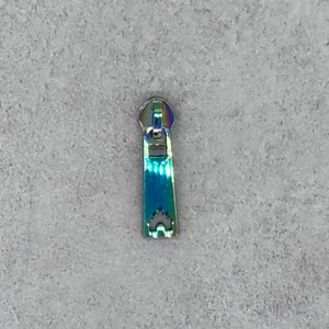 May include: A silver zipper pull with an iridescent finish. The pull has a decorative design and is shaped like a small house.