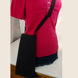 May include: A black fabric crossbody bag with a long adjustable strap. The bag is worn over the shoulder of a person wearing a pink shirt and blue denim shorts.