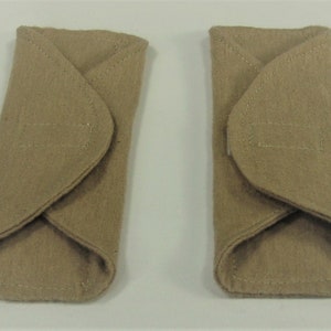 Puede incluir: Dos art&iacute;culos de fieltro beige, de forma rectangular con extremos redondeados. Cada uno tiene una solapa doblada y un detalle rectangular cosido. Se colocan uno al lado del otro sobre un fondo blanco.