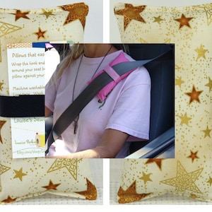 May include: A beige pillow with gold stars printed on it. The pillow is designed to wrap around a car seatbelt to provide comfort and support. The pillow is machine washable. The text on the tag reads "Pillows that ease Wrap the hook and around your seat belt pillow against your shoulder Machine washable Loop tape Wash Louise's Sewing Louise Perkins Etsy.com/shop/LouisePerkins"
