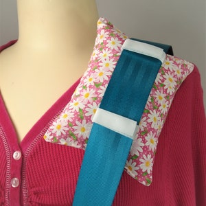 May include: A teal seat belt cover with white accents is placed over a pink cushion with a daisy pattern. The cushion is positioned on a person's shoulder, resting on a pink waffle-knit shirt.
