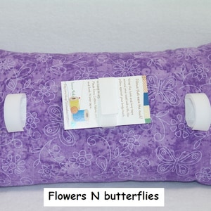 May include: A rectangular purple pillow featuring a white line-art pattern of flowers and butterflies. The pillow includes three white hook-and-loop fastener straps attached to the front. A rectangular product tag is centered on the pillow.