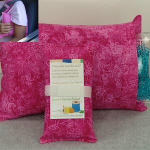 May include: Three pink fabric pillows with white floral designs. The pillows are filled with beads and have a pocket for a cold pack. The text on the tag reads "Pillows that ease the ouch! Wrap the hook and loop taps around your seat belt with the pillow against your body/ouch. Machine washable: Close Hook N. Louise's Sewing Basket. Etsy.com/shop/LouisesSewingBasket"