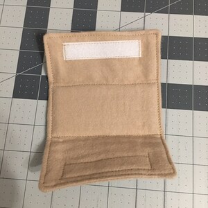 May include: A rectangular tan felt wrap featuring a horizontal white hook-and-loop fastener strip at the top edge and a matching tan hook-and-loop fastener strip positioned along the bottom edge, with visible stitching along all four borders.
