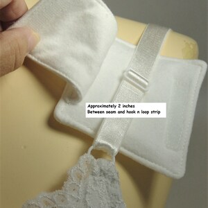 Puede incluir: Una almohadilla de tela blanca para tirante de sujetador con sistema de cierre de velcro. La almohadilla incluye una cinta el&aacute;stica con un regulador de pl&aacute;stico. El texto indica unos 5,08 cm entre la costura y la tira de velcro.