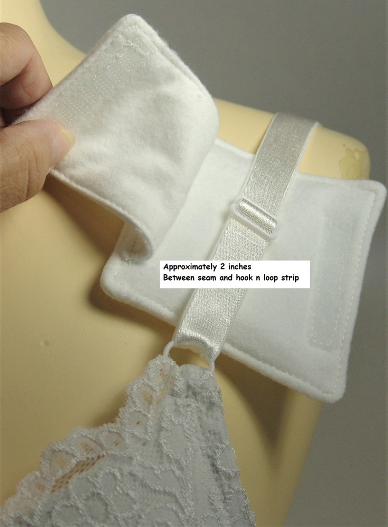 Puede incluir: Una almohadilla de tela blanca para tirante de sujetador con sistema de cierre de velcro. La almohadilla incluye una cinta el&aacute;stica con un regulador de pl&aacute;stico. El texto indica unos 5,08 cm entre la costura y la tira de velcro.