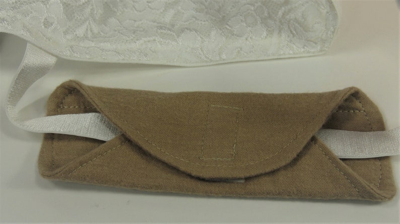 Puede incluir: Un estuche para gafas de fieltro marr&oacute;n con una correa el&aacute;stica blanca. El estuche tiene una solapa curva y est&aacute; cosido con hilo a juego. En el fondo se ve una tela de encaje blanca.