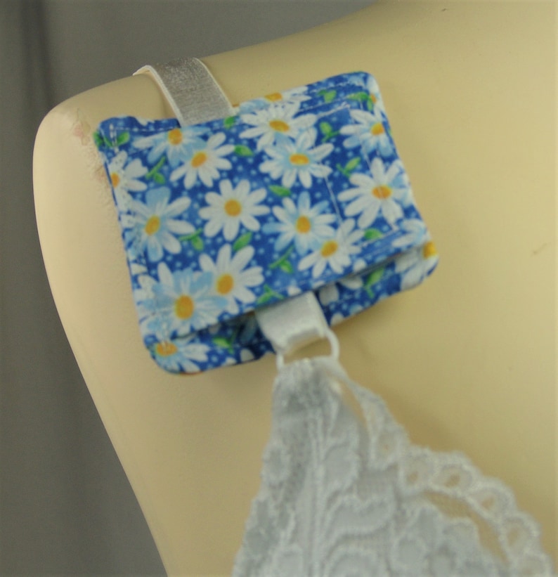 May include: A blue and white daisy-patterned bra strap cover is attached to a white bra strap. The cover is square and made of fabric. The bra strap is attached to a mannequin.
