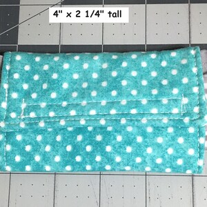 May include: A rectangular teal fabric pouch featuring a white polka dot pattern, attached to a grey and white striped elastic band. The pouch measures 10.16 cm wide by 5.72 cm tall, with visible stitching along the flap and edges.