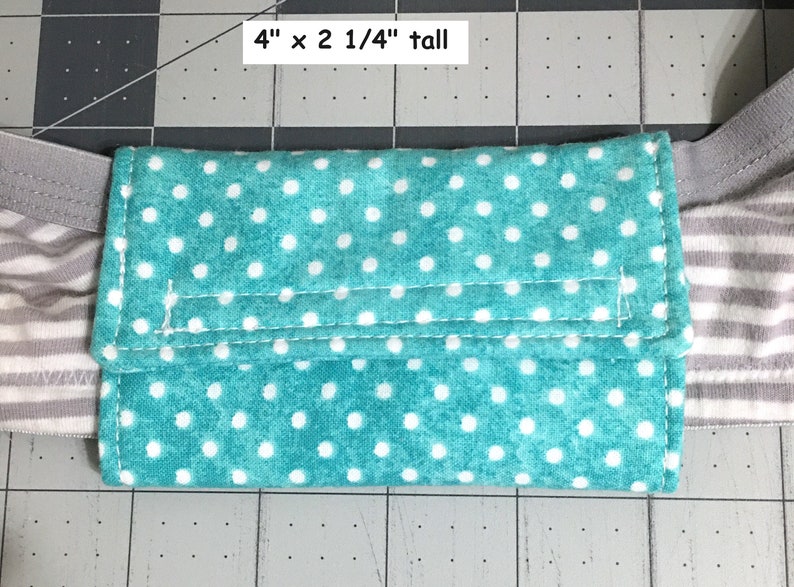 May include: A rectangular teal fabric pouch featuring a white polka dot pattern, attached to a grey and white striped elastic band. The pouch measures 10.16 cm wide by 5.72 cm tall, with visible stitching along the flap and edges.