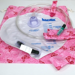 May include: A medical drainage bag with a clear plastic tube and a pink fabric bag and straps. The bag has volume markings and the brand name "Bardia". The pink fabric features a butterfly pattern.