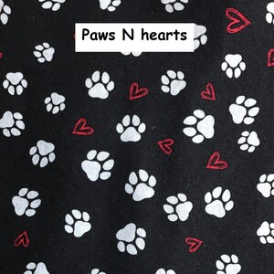Puede incluir: Una tela negra con un patr&oacute;n repetitivo de huellas de animales blancas y peque&ntilde;os corazones rojos delineados. Una etiqueta rectangular blanca con el texto negro Paws N hearts est&aacute; colocada en la parte superior central de la tela.