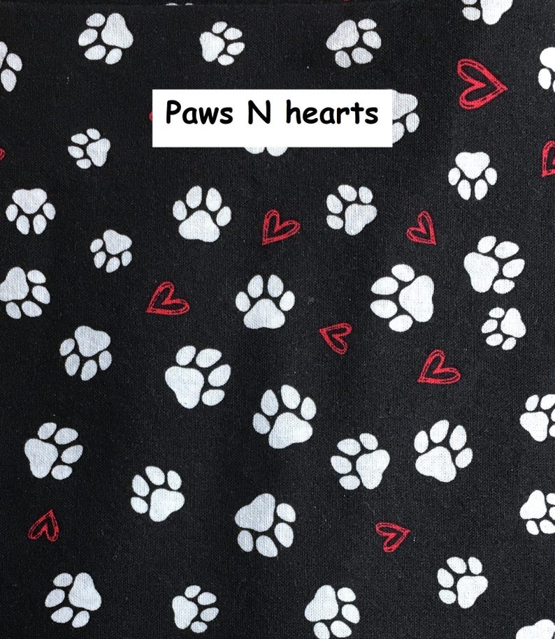 Puede incluir: Una tela negra con un patr&oacute;n repetitivo de huellas de animales blancas y peque&ntilde;os corazones rojos delineados. Una etiqueta rectangular blanca con el texto negro Paws N hearts est&aacute; colocada en la parte superior central de la tela.