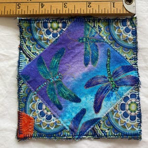 May include: A square fabric coaster with a dragonfly design. The center features a diamond shape with blue and green dragonflies on a purple and blue background. The border has a blue and gold mandala pattern. The coaster measures approximately 3 inches by 3 inches.