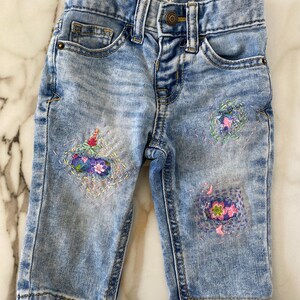 May include: Light blue denim baby jeans with embroidered floral patches in pink, purple, and green. The jeans have a button and zipper closure, front and back pockets, and a faded wash.