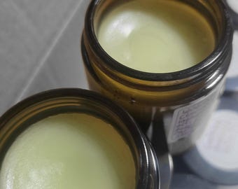Könnte beinhalten: Zwei offene Braunglastiegel mit einem glatten, blassgelben Balsam oder einer Salbe. Jeder Tiegel hat ein weißes Etikett mit aufgedrucktem Text an der Seite. Die Tiegel stehen nah beieinander und zeigen die feste, wachsartige Konsistenz.