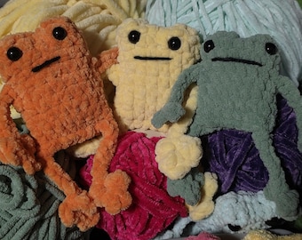 Könnte beinhalten: Drei handgefertigte Frosch-Stofftiere aus weichem, strukturiertem Garn. Die Frösche sind orange, blassgelb und salbeigrün, jeder mit schwarzen Kunststoff-Sicherheitsaugen und einer einfachen, gestickten schwarzen Linie als Mund sowie langen Gliedmaßen.