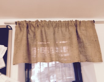 May include: A brown burlap valance hangs from a metal rod above a window. The valance is gathered in the center and has a simple, rustic look.