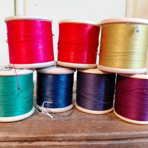May include: A selection of colourful thread spools in shades of red, teal, navy, purple, and gold. The thread is wound around wooden spools, creating a visually appealing display. The spools are arranged on a wooden surface.