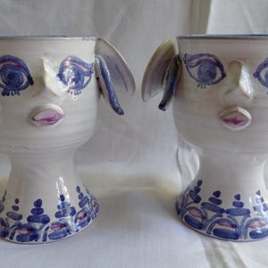 May include: Two quirky ceramic face planters. Each white planter has blue and purple painted details, including eyes, lips, and decorative patterns. The planters have ear-like handles.