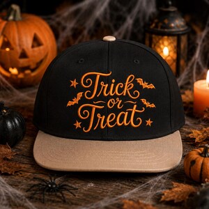 May include: A black baseball cap with a tan brim featuring orange embroidered text that reads Trick or Treat, accented with small orange stars and flying bat silhouettes. The cap has a small tan button on the top centre of the crown.