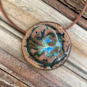 May include: A brown ceramic pendant with a blue and green glaze. The pendant is round and has a textured surface. It is hanging from a brown cord.