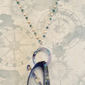 May include: A teal and white beaded chain with a clear plastic ring for holding glasses. The chain is hanging on a map-like background.